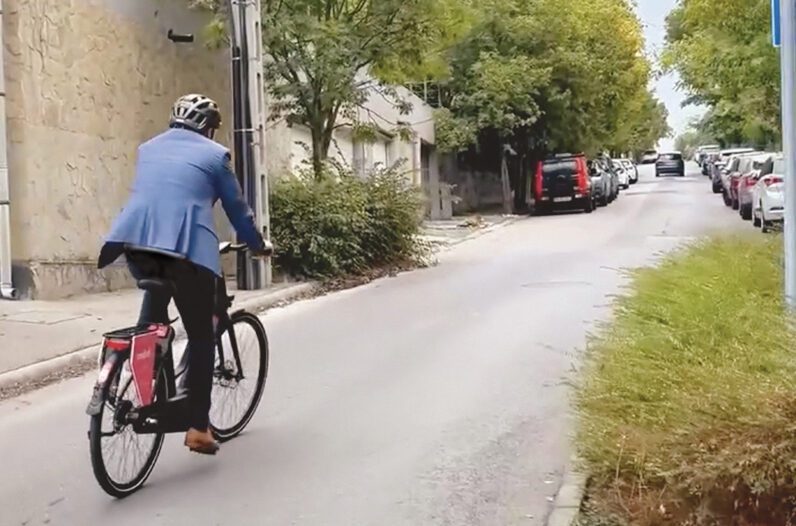 Egy bukósisakos, öltönyös férfi ekerékpárral távolodik egy városi betonúton.