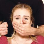 Egy nő mögött álló férfi bal kezével befogja egy nő száját, jobb kezét pedig ökölbe szorítja.