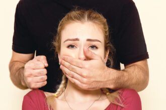 Egy nő mögött álló férfi bal kezével befogja egy nő száját, jobb kezét pedig ökölbe szorítja.