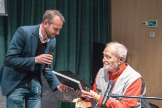 Dr. Juhász Árpád éppen egy keretezett képet vesz a kezébe, melyet vendéglátója ad át neki a beszélgetés során.