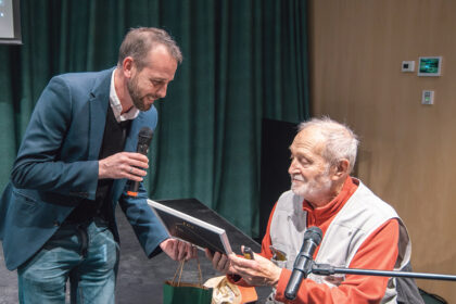 Dr. Juhász Árpád éppen egy keretezett képet vesz a kezébe, melyet vendéglátója ad át neki a beszélgetés során.